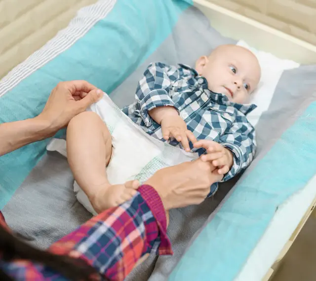 Mama sprawdza, kiedy zmienić rozmiar pieluchy maluszkowi. Dziecko leży na przewijaku w kratkę.
