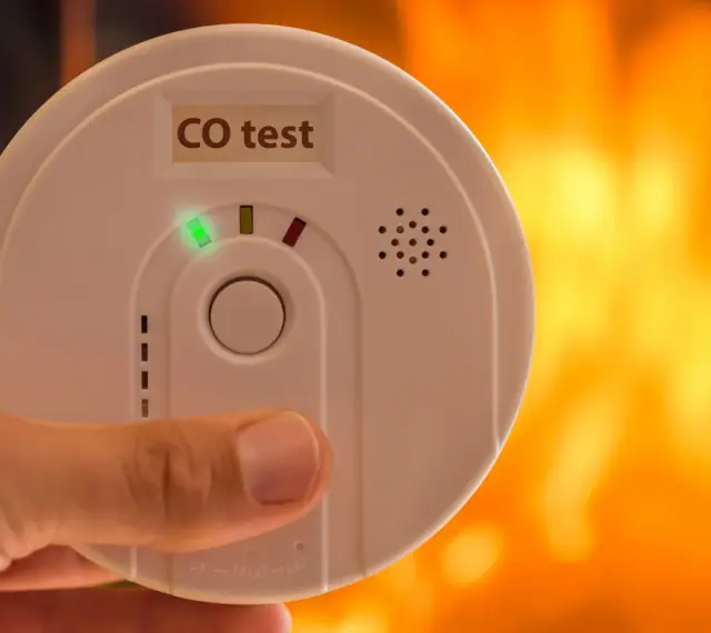 Detektor czadu z zieloną diodą i napisem "CO test" na tle płomieni.