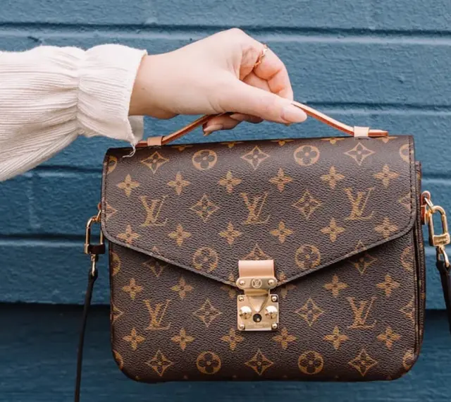 Una mano tiene una borsa Louis Vuitton con il classico monogramma. Un'icona di stile, che ci ricorda dove louis vuitton nasce: dalla maestria artigianale francese.