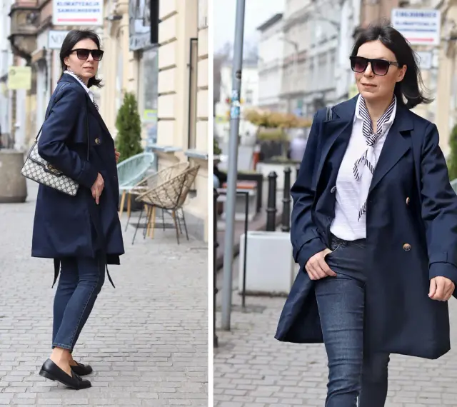 Elegancka kobieta w granatowym trenczu damskim, białej bluzce w paski i jeansach spaceruje po brukowanej ulicy.