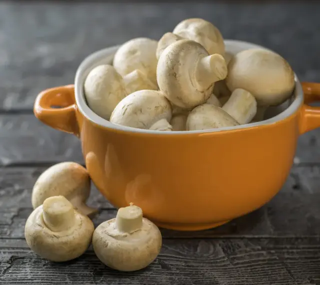 Pomarańczowa miseczka pełna świeżych pieczarek, jakby w ciąży z nowymi smakami. Kilka pieczarek leży obok na drewnianym blacie.
