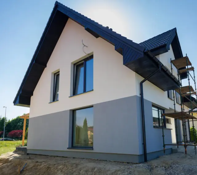 Nowy dom z dwukolorową elewacją, biało-szarą, to świetny pomysł na elewację domu. Widać rusztowania, co sugeruje trwające prace.