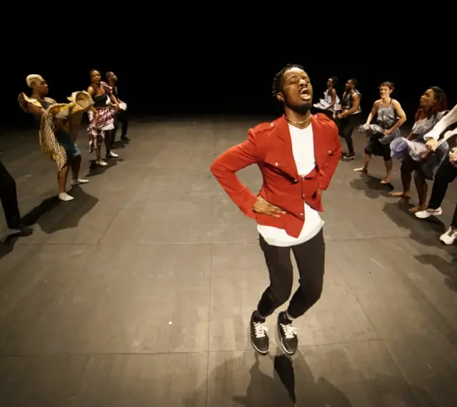 Un danseur afro 93, en veste rouge, mène une troupe sur scène. Les danseurs, aux tenues variées, exécutent des mouvements synchronisés.
