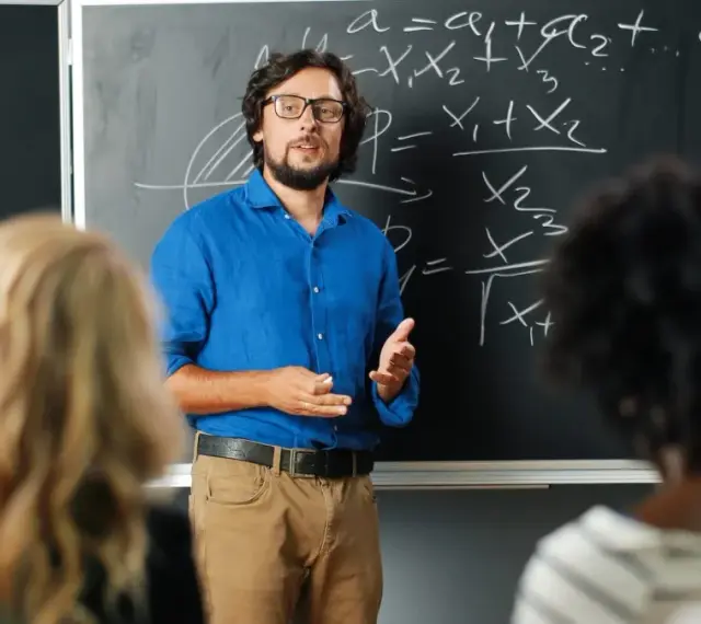 Nauczyciel w niebieskiej koszuli tłumaczy matematykę przy tablicy. Czy nauczyciel jest funkcjonariuszem publicznym? To pytanie nurtuje wielu.
