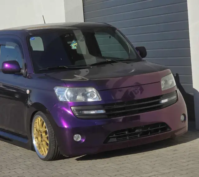 Fioletowy Daihatsu Materia z błyszczącymi złotymi felgami, zaparkowany przed garażem.