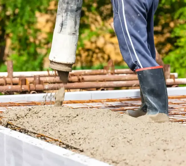 Beton B30 wylewany z pompy na zbrojenie. Pracownik w gumowcach stoi w świeżej masie, pilnując prawidłowych proporcji.