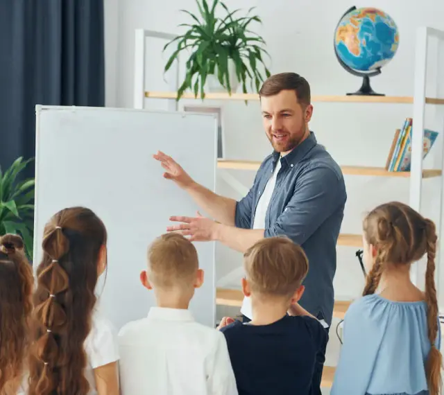 Nauczyciel tłumaczy coś dzieciom przy tablicy. To mogą być dni dyrektorskie, podczas których uczniowie poznają szkołę.