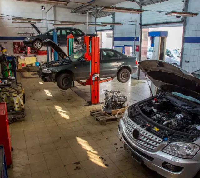 Warsztat samochodowy z kilkoma autami na podnośnikach, w tym ciemnym sedanem, który wygląda jak p1351 peugeot. Silnik wyjęty z jednego z pojazdów.