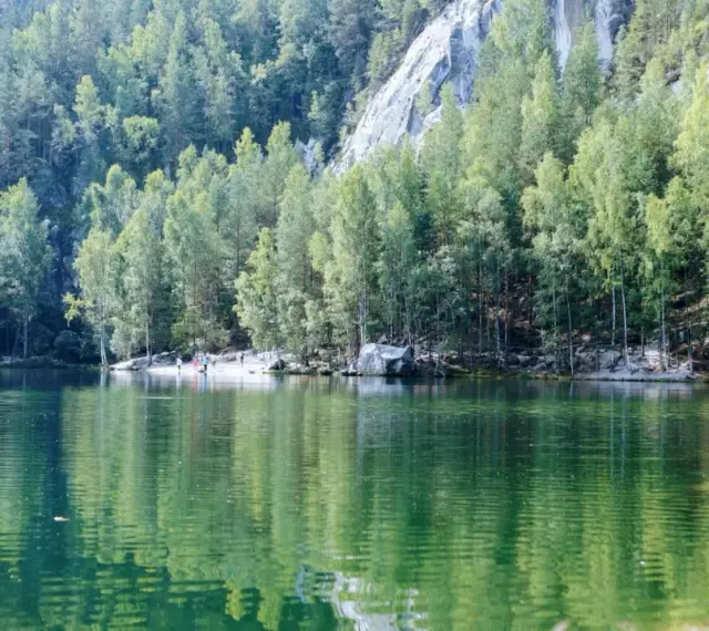 Zielona tafla jeziora odbija las i skalne miasto w Polsce. Ludzie spacerują po piaszczystym brzegu.