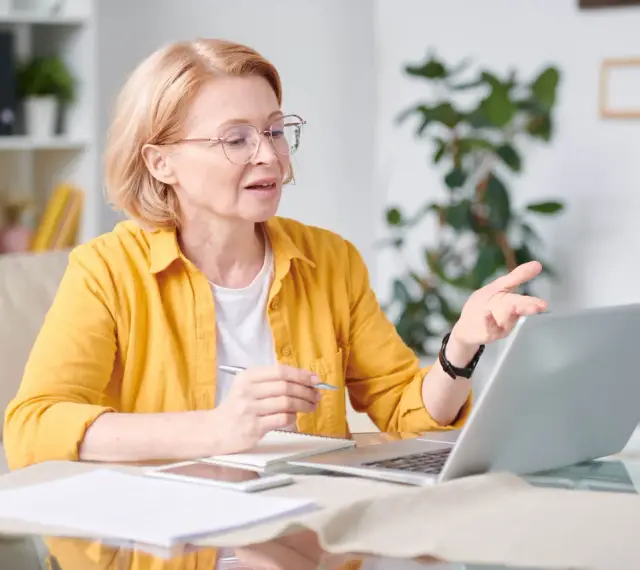 Kobieta w okularach i żółtej koszuli pracuje przy laptopie, trzymając długopis. To jej dodatek funkcyjny do pracy zdalnej.