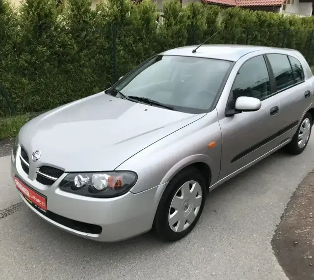 Srebrny Nissan Almera hatchback stoi na asfaltowej drodze, otoczony zielenią.
