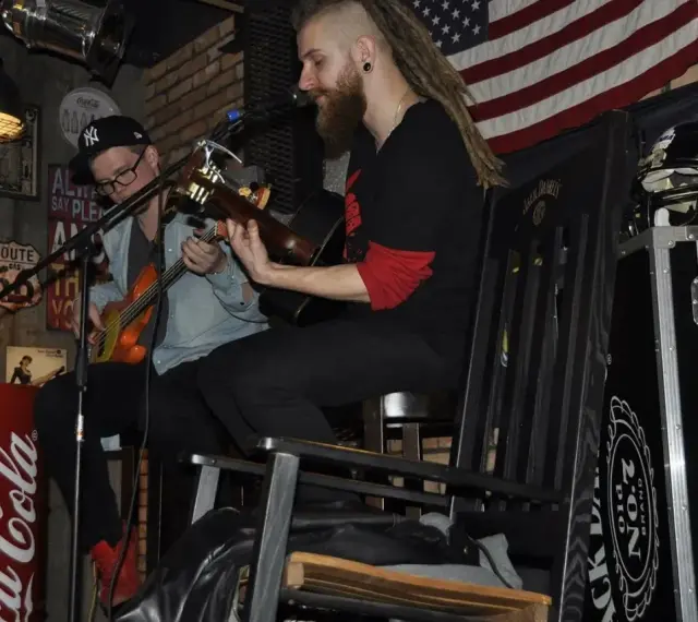 Dwóch muzyków gra na gitarach w klimatycznym wnętrzu. Jeden z nich, muzyk grający do kotleta, ma dredy i brodę.