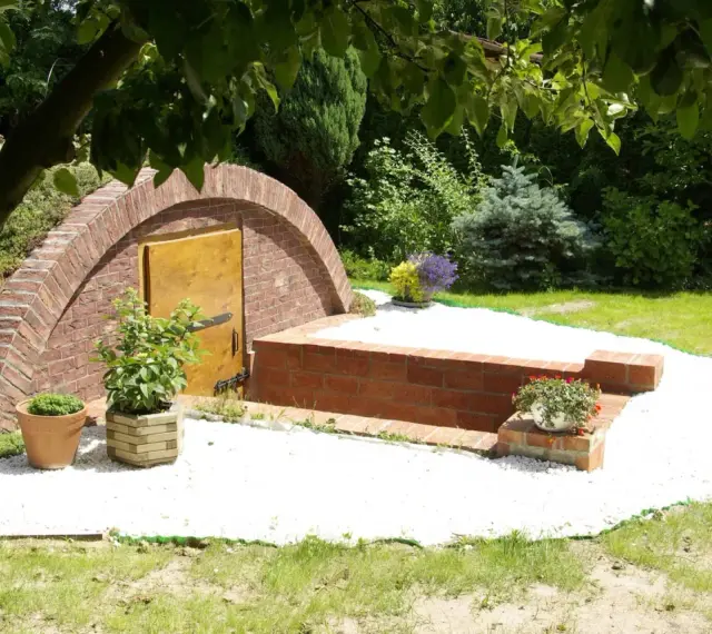 Ziemianka z cegły z drewnianymi drzwiami, otoczona białym żwirem i zielenią. Zastanawiasz się, ile kosztuje ziemianka?