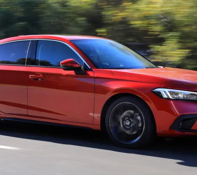 Czerwony Honda Civic 11. generacji pędzi po drodze, otoczony zielenią drzew.