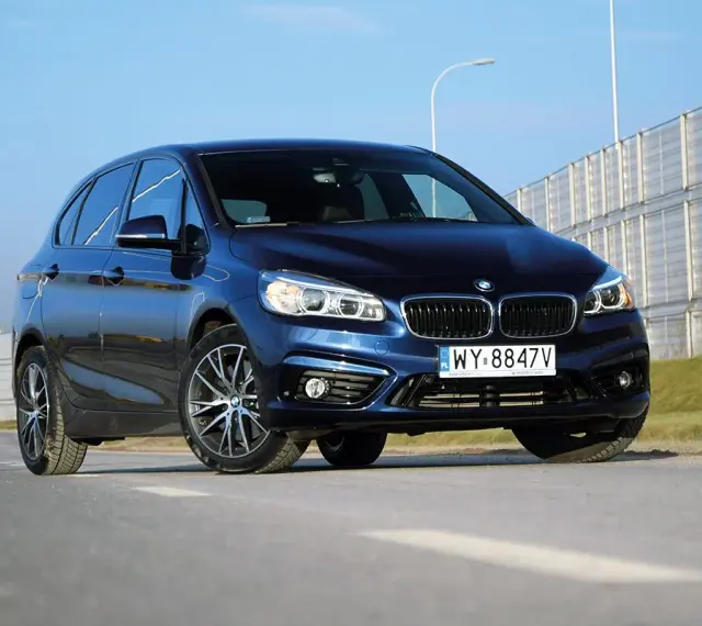 Ciemnoniebieski BMW van na tle jasnego nieba i barier dźwiękochłonnych.
