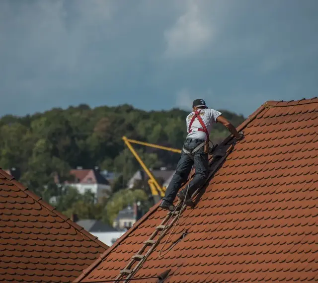 Robotnik w kasku i szelkach bezpieczeństwa na dachu, pracujący przy dachówce.