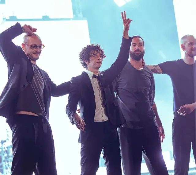 Matty Healy i zespół The 1975 na scenie. Muzycy pozdrawiają publiczność, jeden z nich unosi rękę w geście pokoju.