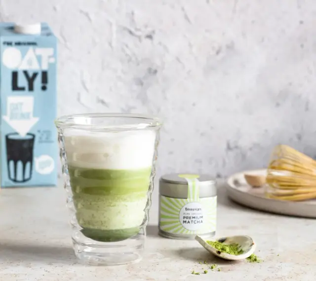 Orzeźwiający iced matcha latte z pianką, obok puszka matcha i bambusowa miotełka. Idealny napój na ciepłe dni.