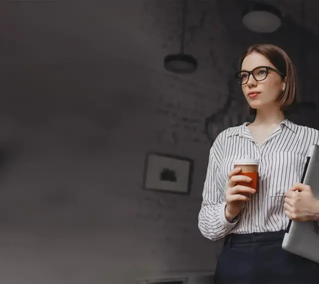 Kobieta w okularach trzymająca kawę i laptopa.