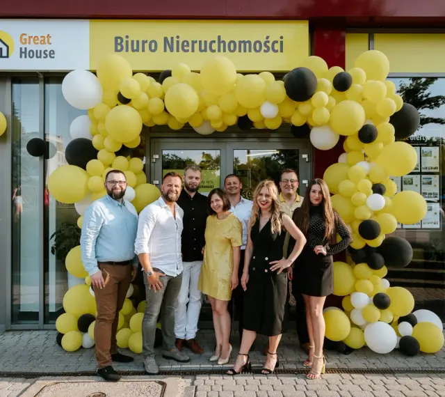 Grupa ludzi pozuje przed biurem nieruchomości ozdobionym żółtymi balonami.