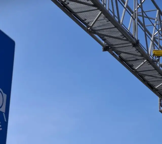 Niebieski znak informuje o odcinkowym pomiarze prędkości S7. Nad drogą widać konstrukcję z żółtą kamerą.