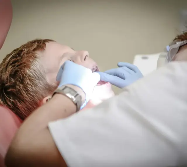 Chłopiec siedzi na fotelu dentystycznym podczas wizyty u dentysty. Dentysta w rękawiczkach bada jego zęby.