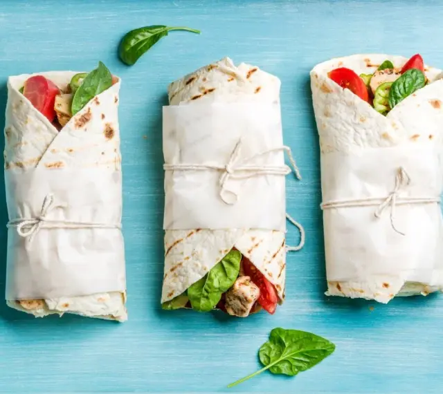 Trzy keto tortille z siemienia lnianego, zawinięte w papier i przewiązane sznurkiem, z widocznym nadzieniem z kurczaka, pomidorów i szpinaku.