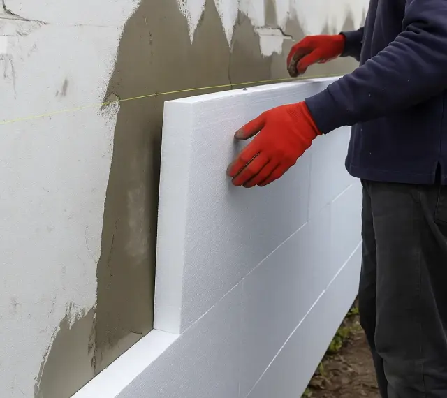 Pracownik w czerwonych rękawicach montuje styropian na ścianie. Dowiedz się, ile kosztuje elewacja domu.