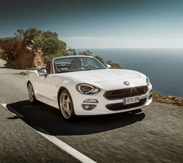 Biały Fiat 124 Spider sunie po krętej drodze nad morzem.