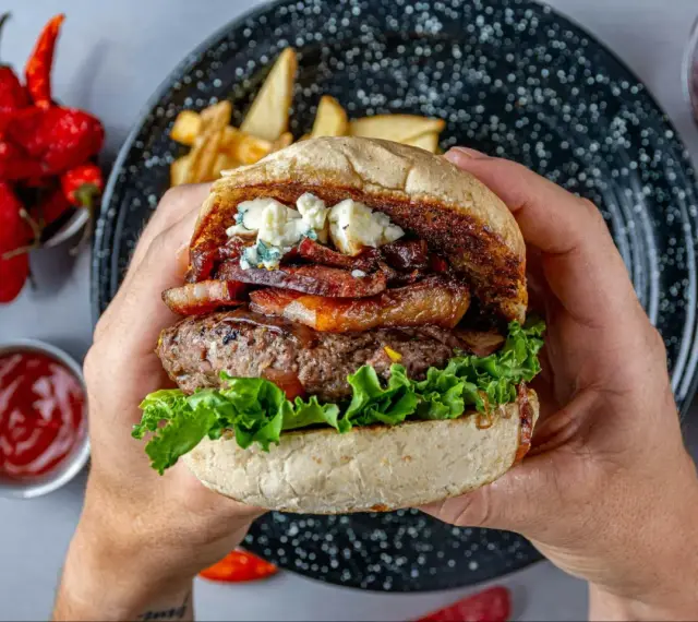 Soczysty burger wołowy z serem pleśniowym, boczkiem i sałatą, trzymany w dłoniach.