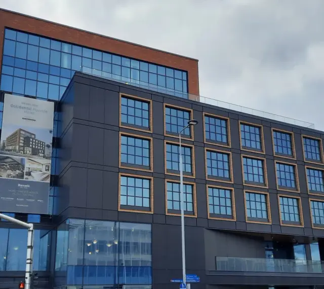 Nowoczesny budynek z cegły i ciemnej fasady, z banerem zapowiadającym otwarcie Occidental Powisle Hotel, części sieci Barceló.