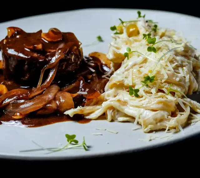 Mięso w sosie z cebulą i grzybami, przypominającymi podgrzybki, podane z kremowym makaronem posypanym parmezanem i kiełkami.