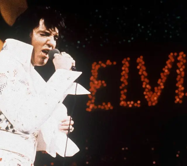 Elvis Presley singt in seinem berühmten weißen Anzug in Las Vegas, während sein Name in roten Lichtern erstrahlt.