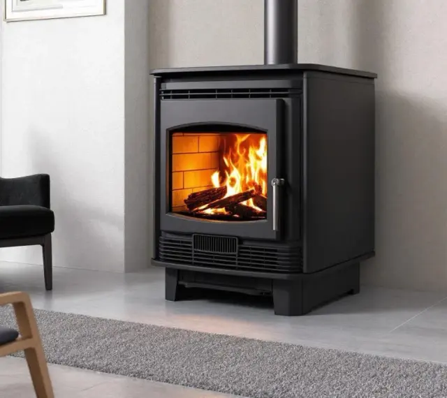 Poêle à bois moderne, flammes vives réchauffant la pièce. Les granulés ne tombent plus, juste du bois qui crépite. Un fauteuil noir et une plante verte complètent la scène.