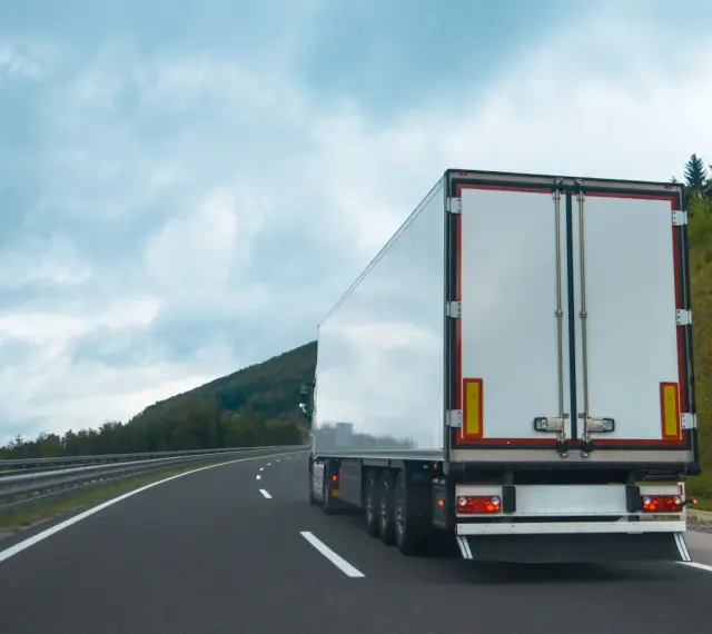 Ciężarówka na autostradzie, z tyłu widoczne białe drzwi naczepy. W tle góry i chmury. Tablice wyróżniające przepisy są częścią oznakowania pojazdu.