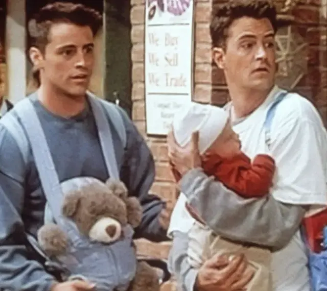 Chandler i Joey z dzieckiem i misiem w nosidełku. Chandler wygląda na lekko zaniepokojonego.
