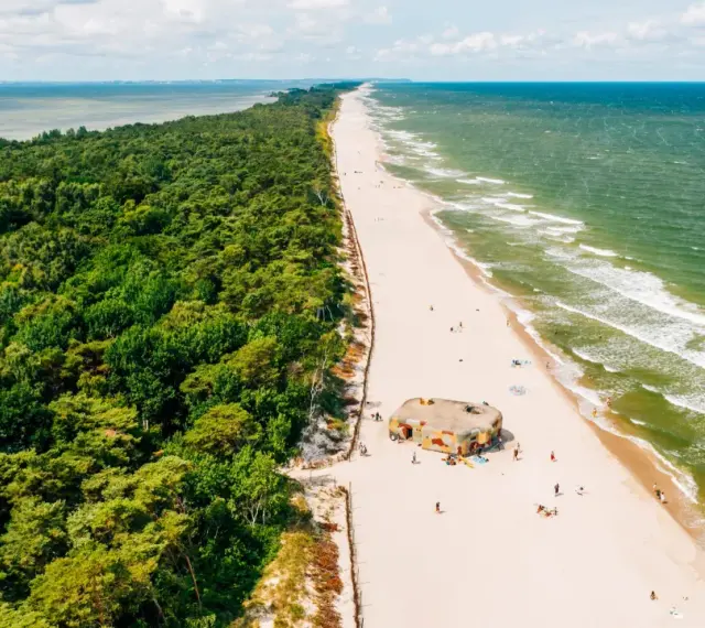 Majówka nad morzem: zielony las, piaszczysta plaża z kilkoma osobami i bunkrem w kamuflażu, szmaragdowe fale Bałtyku.