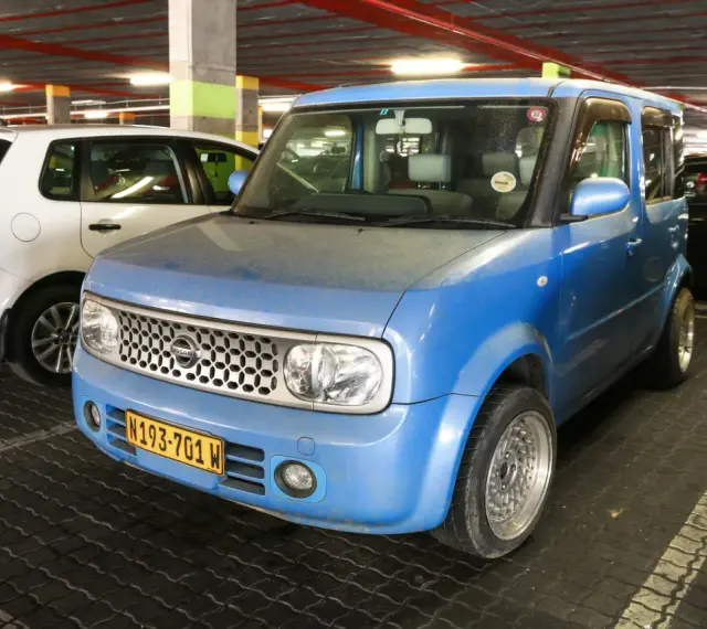 Niebieski Nissan Cube z charakterystycznym grillem i tablicą rejestracyjną N193-701 W, zaparkowany obok białego Volkswagena.
