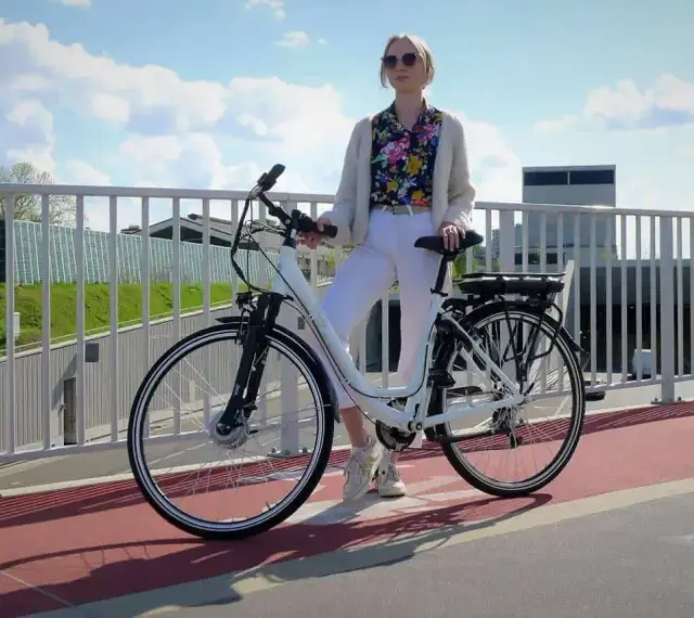 Kobieta w białych spodniach i kolorowej koszuli stoi obok białego roweru elektrycznego. Czy na rower elektryczny trzeba mieć prawo jazdy?