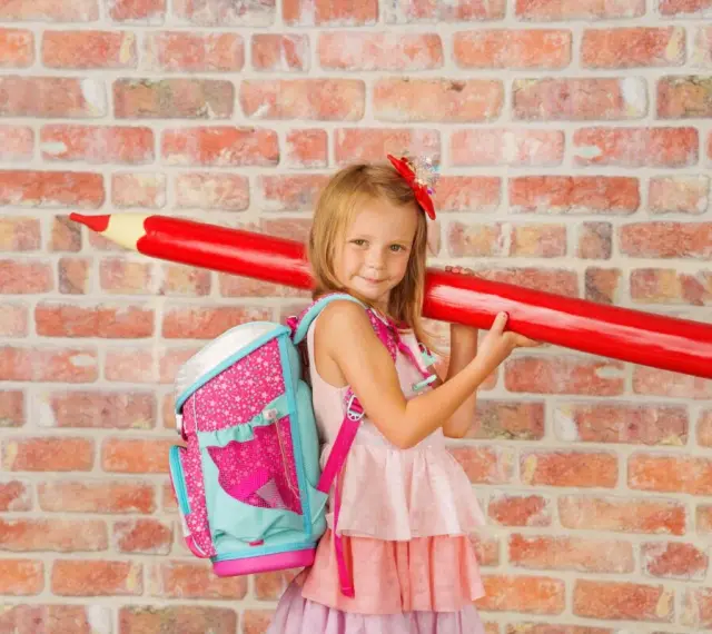 Ein Mädchen mit pinkem Rucksack und riesigem roten Stift vor Ziegelwand. Bereit für die Einschulung Berlin 2025!