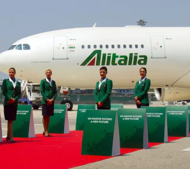 Cztery stewardessy w zielonych uniformach stoją na czerwonym dywanie przed samolotem Alitalia.