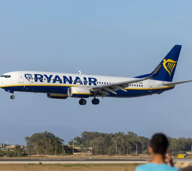 Samolot Ryanair lądujący na tle nieba i drzew, z widoczną sylwetką osoby w pierwszym planie.
