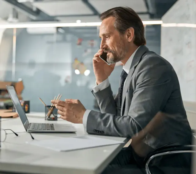 Biznesmen w garniturze rozmawia przez telefon, pracując przy laptopie. To typowy dzień w życiu CEO, co to znaczy być liderem.
