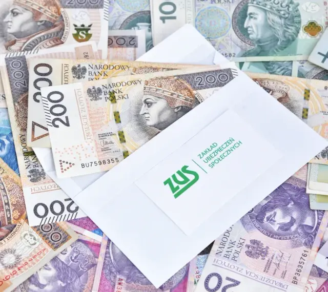Koperta ZUS na stosie polskich banknotów. Może to być symbol wypłaty zasiłku przedemerytalnego lub innych świadczeń.