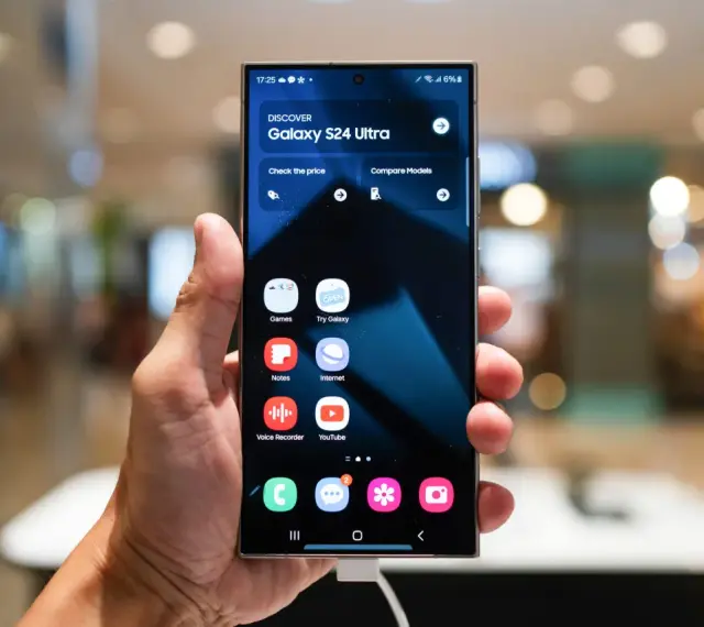 Dłoń trzyma smartfon z interfejsem One UI 6, prezentującym ekran startowy Galaxy S24 Ultra z ikonami aplikacji.