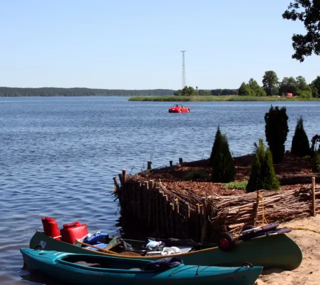 Dwie kajaki na piaszczystej plaży w Swornegaciach na Kaszubach. W oddali czerwona łódka i jezioro.
