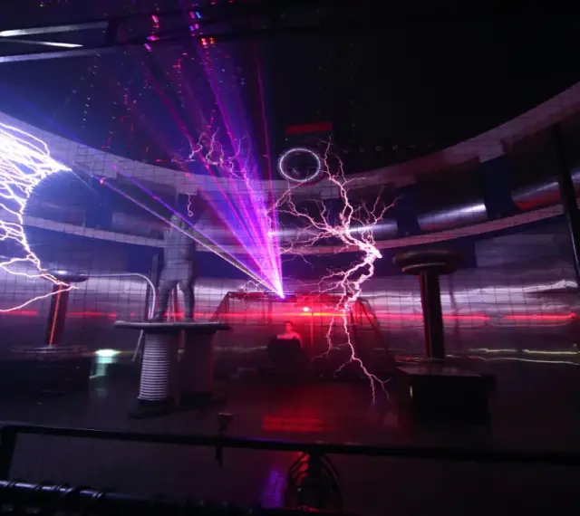 Spektakuläre Blitze zucken in einem **Faradayschen Käfig**, während ein Mann sicher darin steht. Laserstrahlen durchschneiden die Dunkelheit.