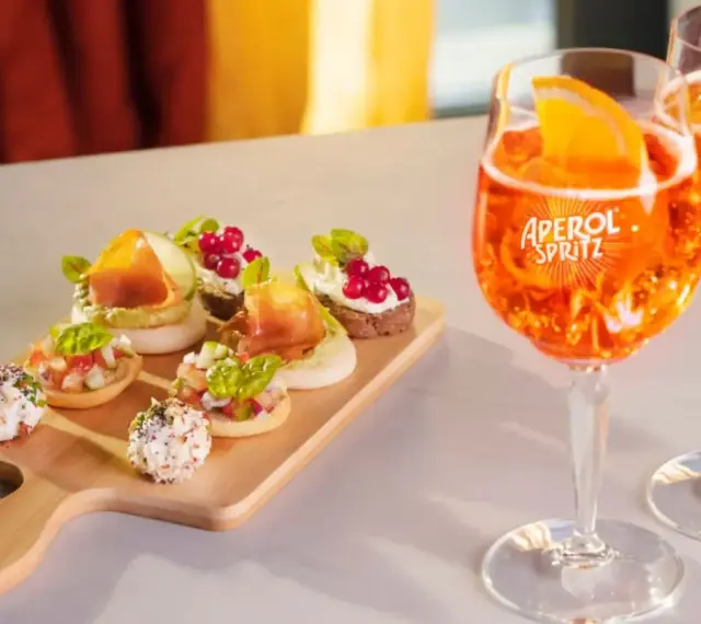 Zwei Aperol Spritz Gläser mit Orangenscheiben und eine Holzplatte mit verschiedenen Häppchen. Perfekt für einen Aperitif!