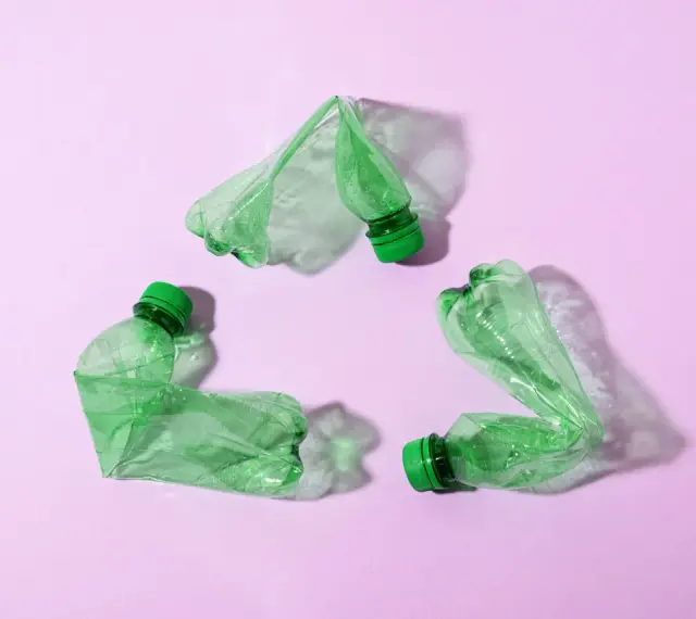 Trzy zielone, zgniecione butelki tworzą symbol recyklingu na różowym tle.