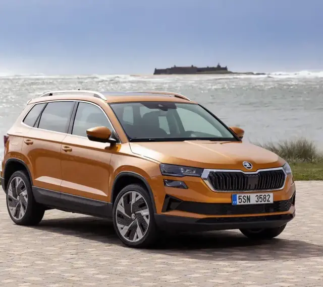 Pomarańczowa Skoda Karoq na tle morza i zamku. Sprawdź aktualny skoda cennik i wybierz swój wymarzony model.